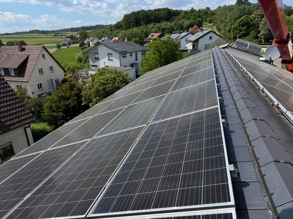 Beispielbild eines Hausdachs und den installierten PV Paneelen mit einem Dorf im Hintergrund. Panoramablick mit den 48 sichtbaren Paneelen, was etwa 22 kWP Leistung ergibt. Aufgenommen vom Plateau eines Krans